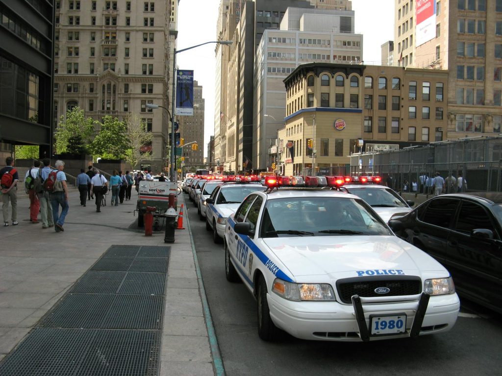 A line of NYPD police cars with flashing lights on a busy New York street, featuring pedestrians and tall buildings.