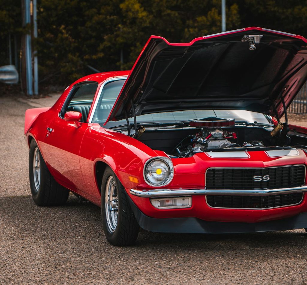 A striking red vintage Camaro with its hood open, showcasing the engine on a city street.