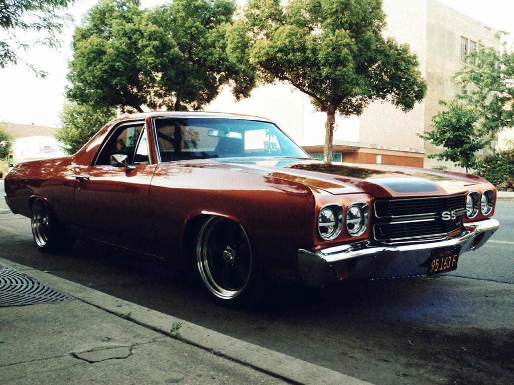Red Chevrolet El Camino parked on a tree-lined city street, evoking a nostalgic and retro feel.