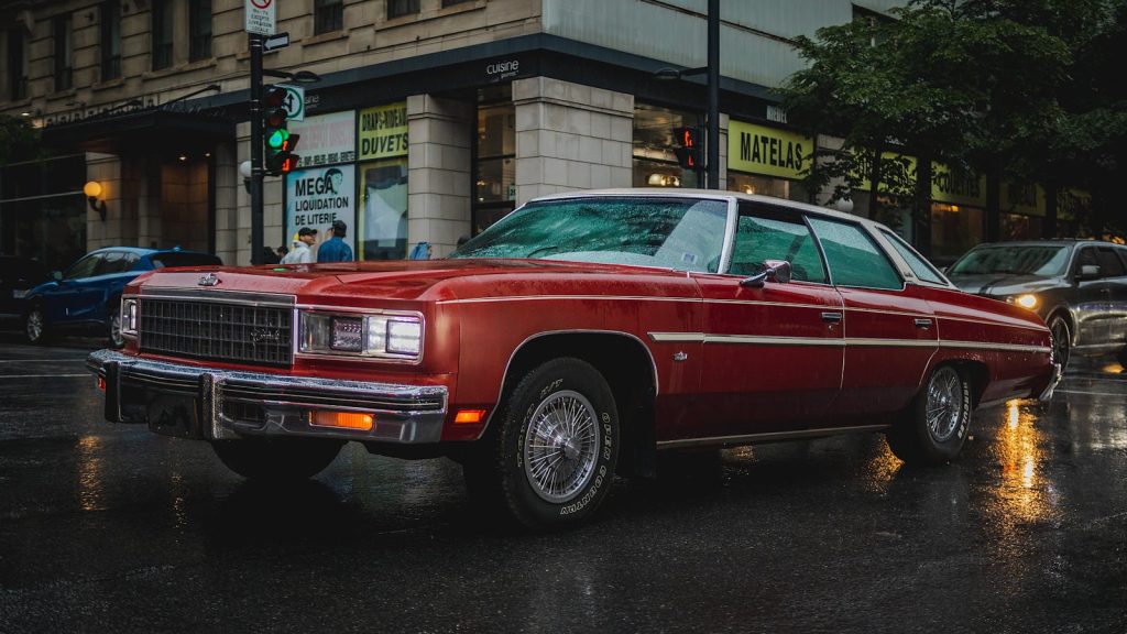 A red vintage car parked on a rainy city street, reflecting urban nostalgia.