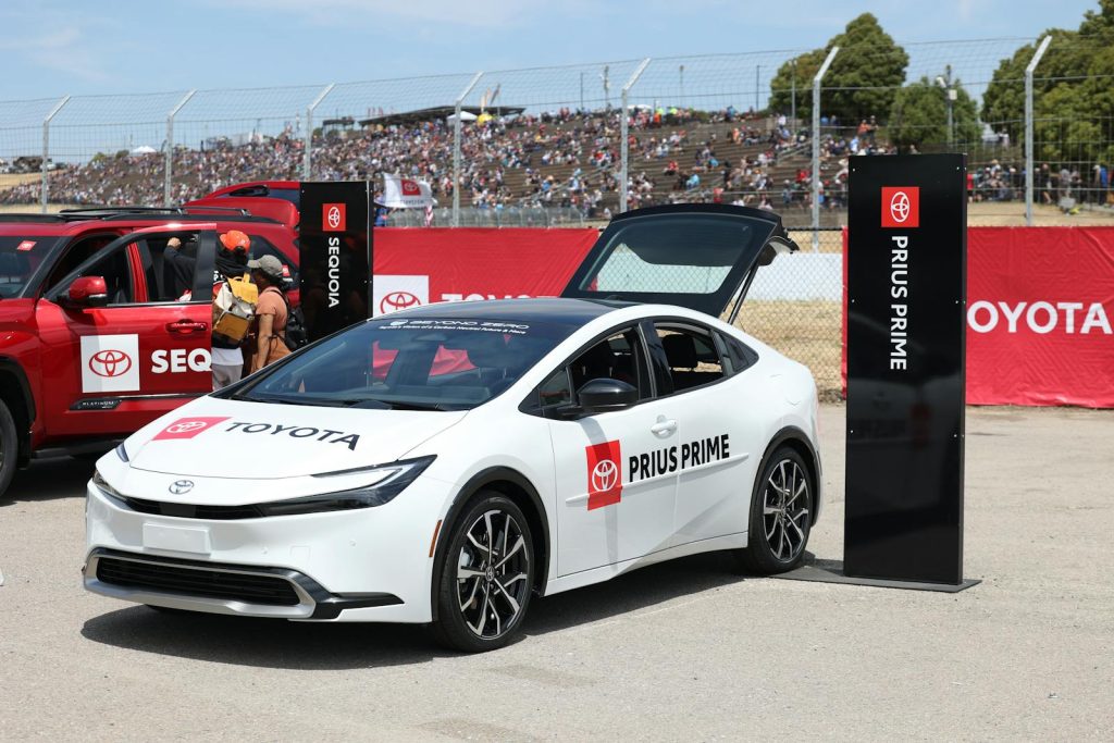 A Toyota Prius Prime on display at an outdoor car exhibition with spectators in the background.