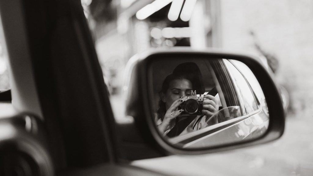 Black and white photo of a woman photographing through a car side mirror.