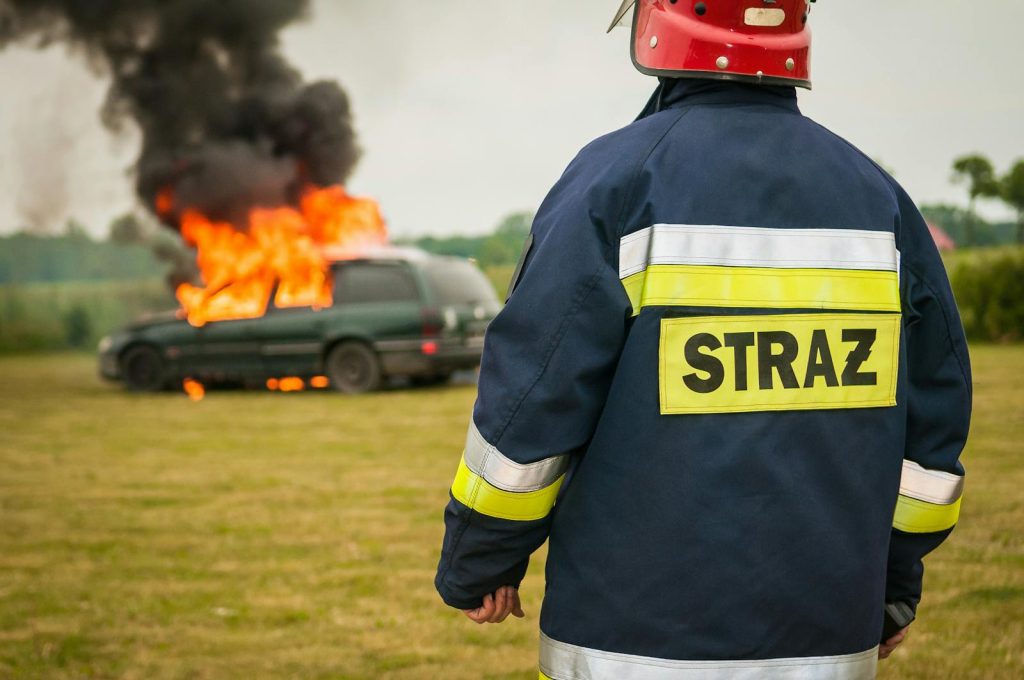 Firefighter observing a burning car with thick black smoke outdoors.