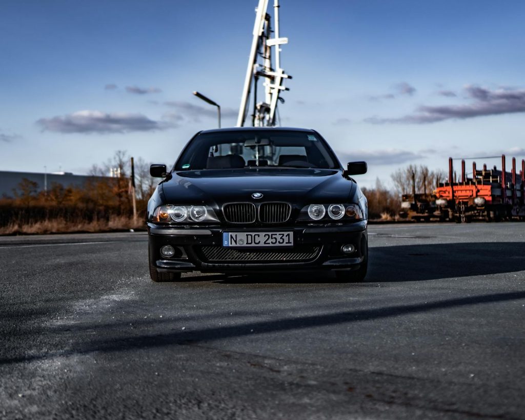 Front view of a sleek black BMW E39 car parked in an industrial area under a clear sky.