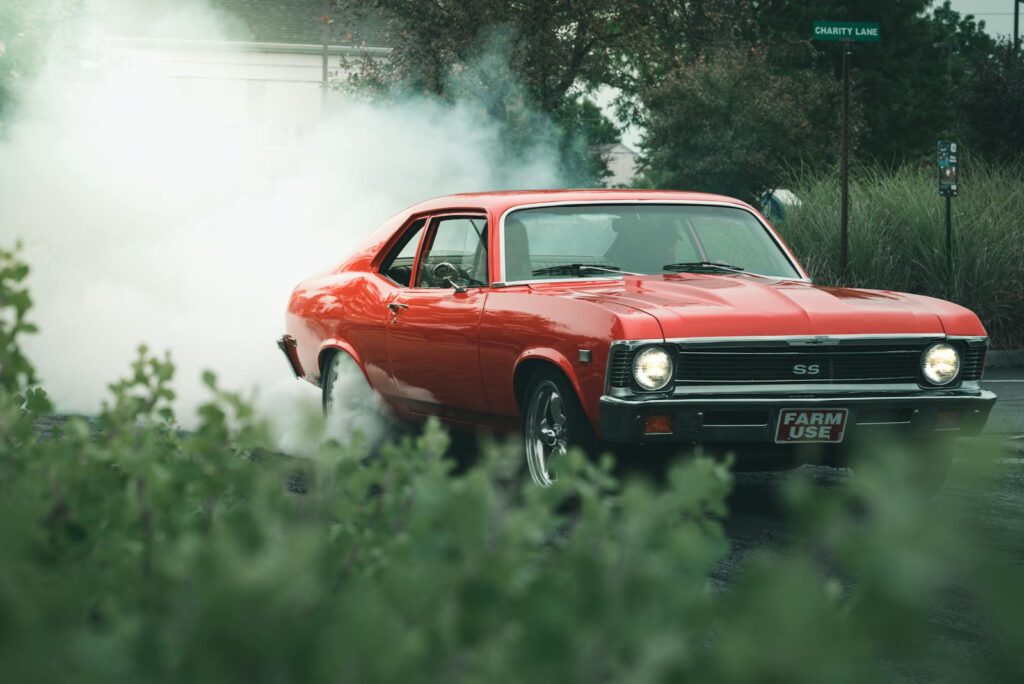 A classic orange muscle car doing a burnout on a street with smoke drifting around.