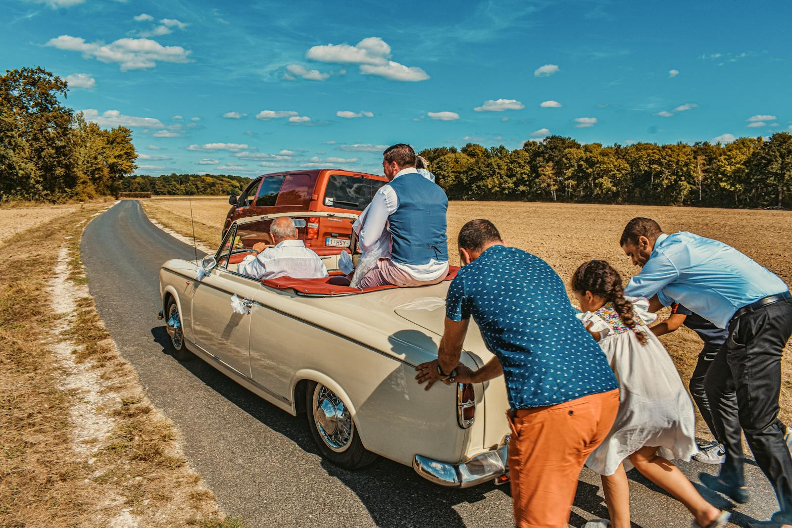 Family pushes classic wedding car in countryside, a humorous moment on sunny day.