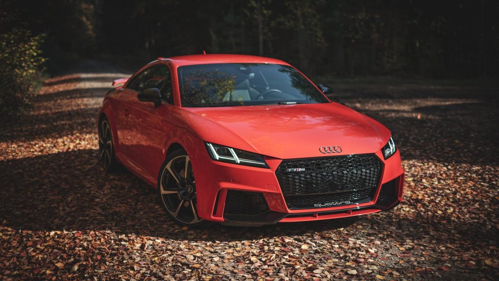 Vibrant red sports car parked on a leaf-strewn road in a forest setting captured in daylight.