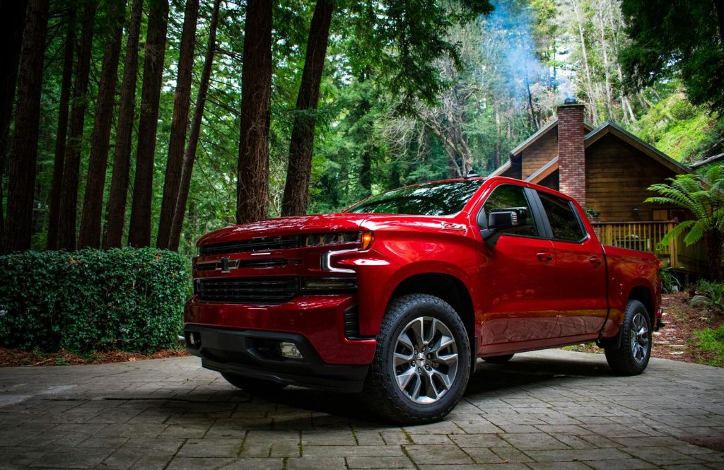 A red Chevrolet Silverado 1500 parked in a rustic driveway surrounded by lush forest and cottage.
