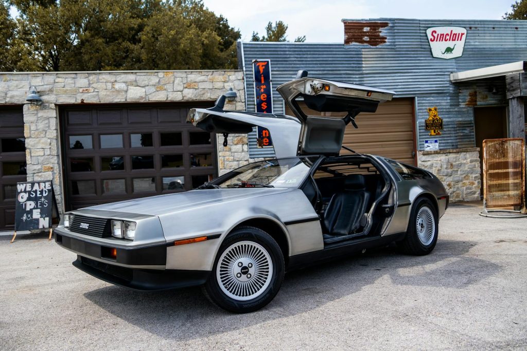 A classic DeLorean DMC-12 with gullwing doors open, parked at a retro Sinclair garage.