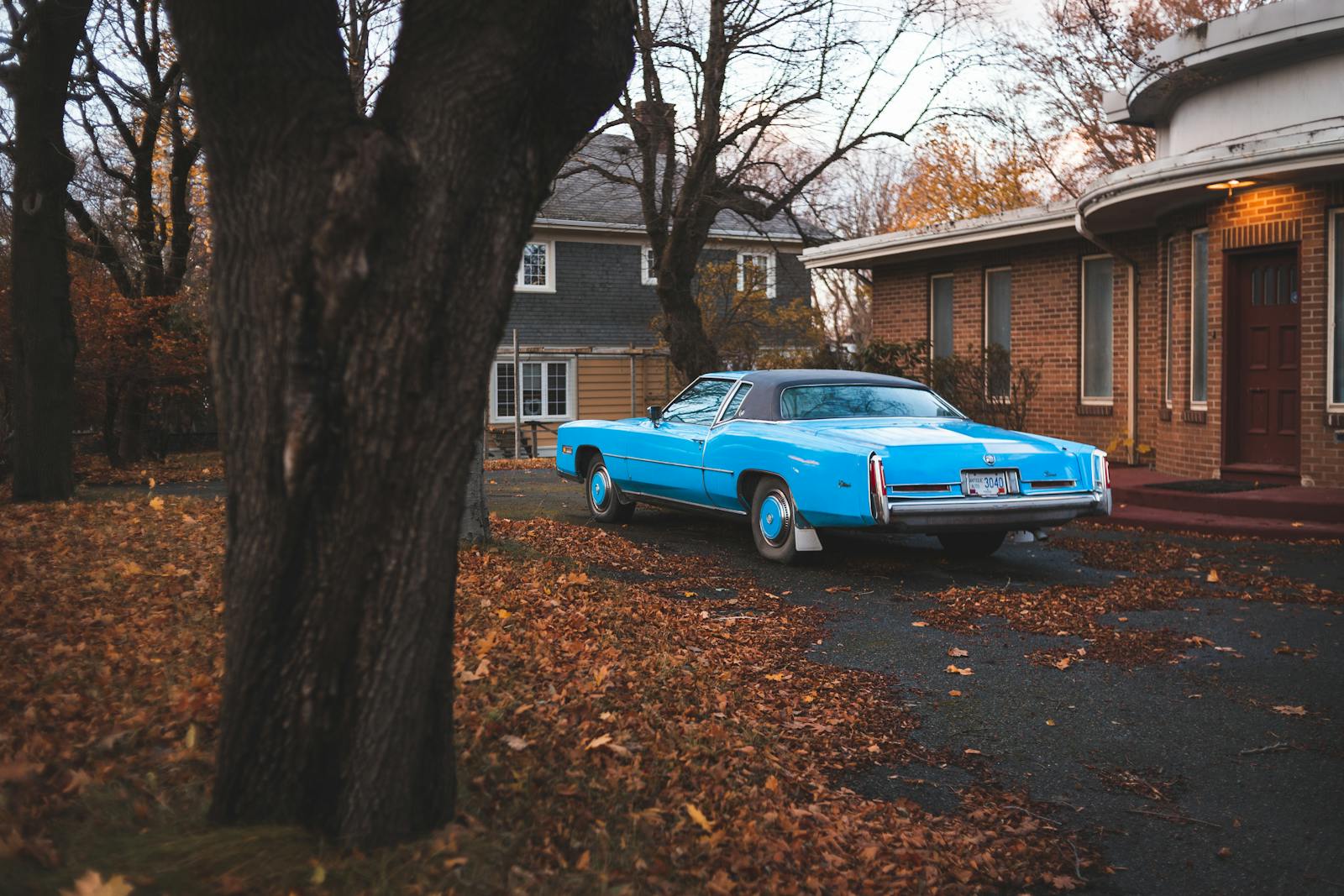 A classic blue car parked on a leaf-covered driveway beside a brick house during autumn.