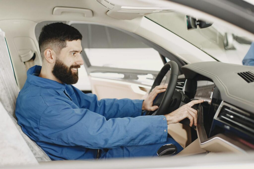 Mechanic in blue coveralls interacts with car dashboard, smiling and focused.