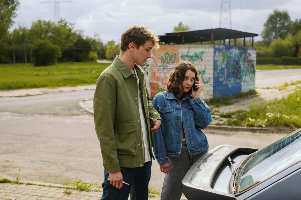A young couple stands by their car, possibly facing a breakdown.