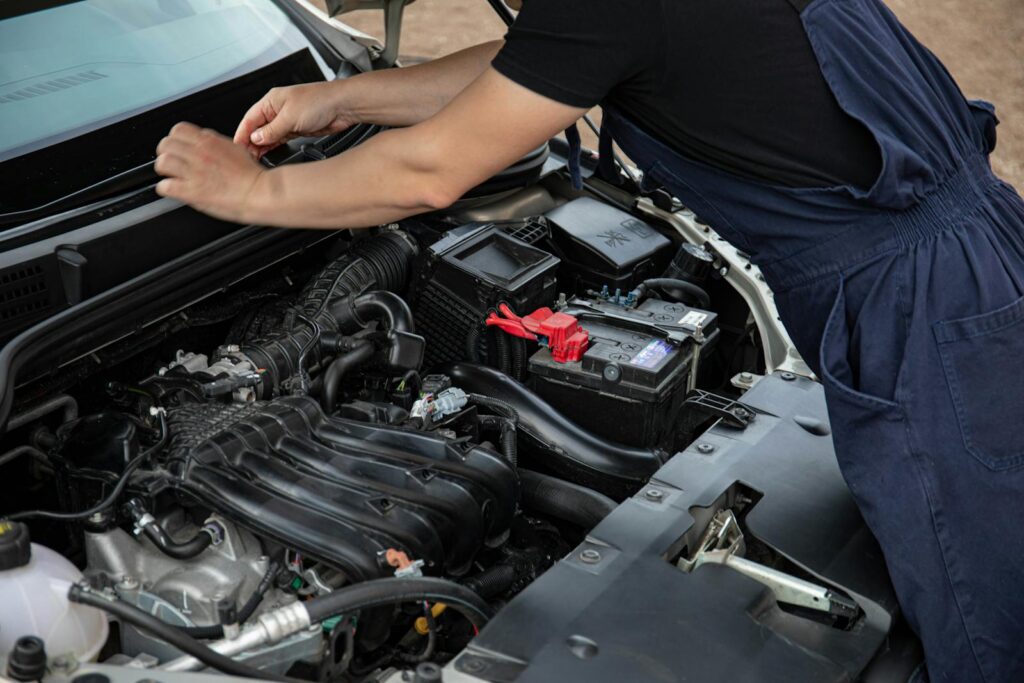 Mechanic performing maintenance on a car engine outdoors during the day.
