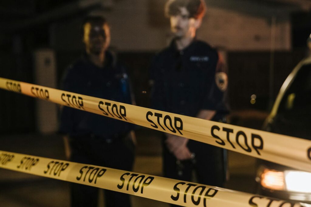 Two police officers standing behind a stop tape at night, scene partially illuminated by car headlights.