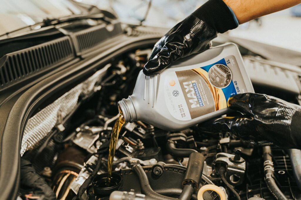 A mechanic wearing gloves pouring oil into a car engine in a garage setting.