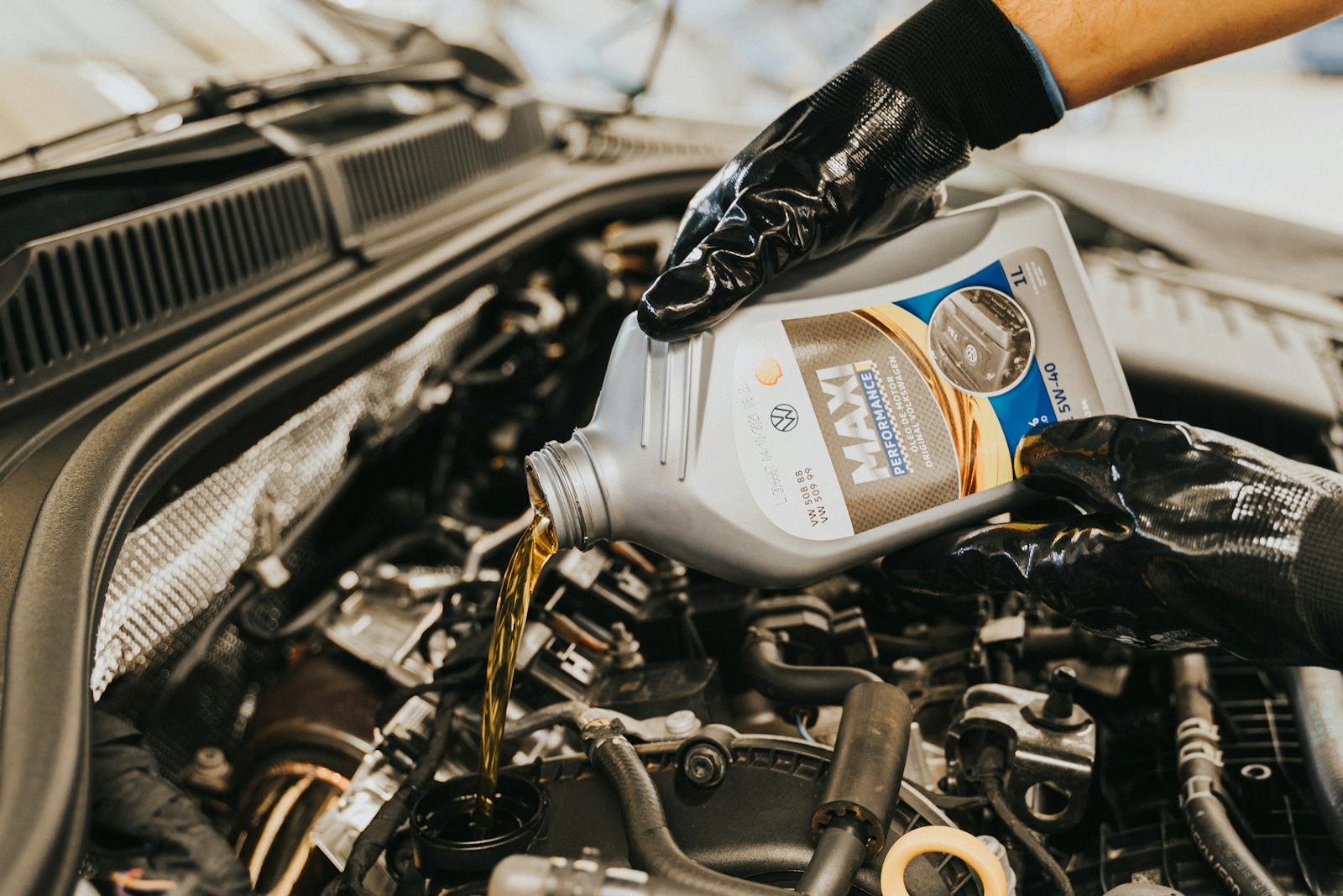 A mechanic wearing gloves pouring oil into a car engine in a garage setting.