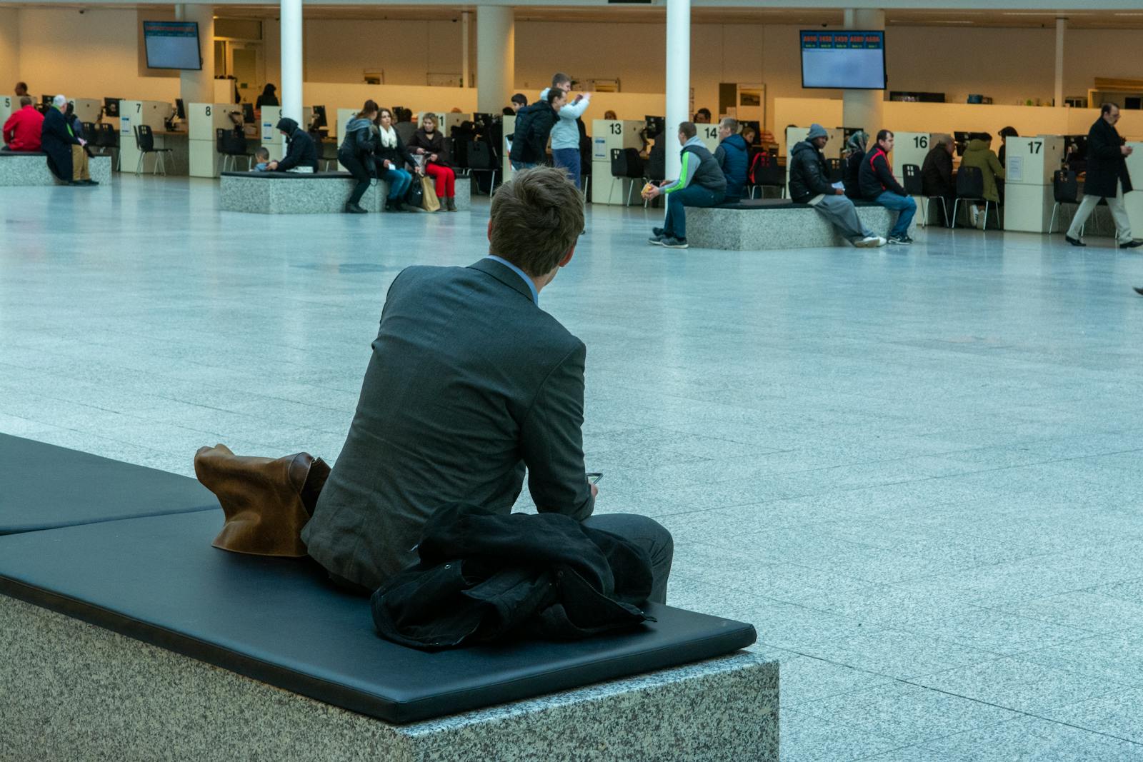 Businessman in suit seated in airport terminal waiting area, observing travelers.