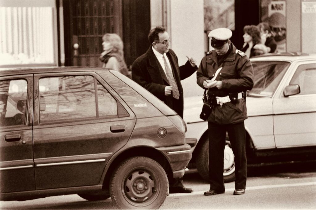 A police officer issues a parking ticket to a driver on a busy city street. Sepia-toned.