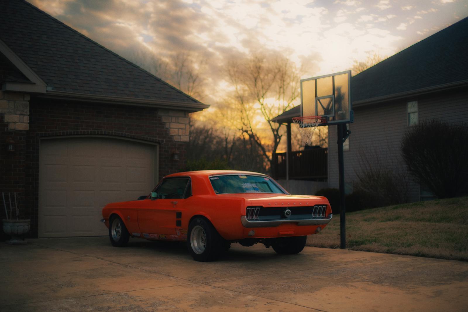 A vintage red muscle car parked in a tranquil village driveway under a beautiful sunset sky.