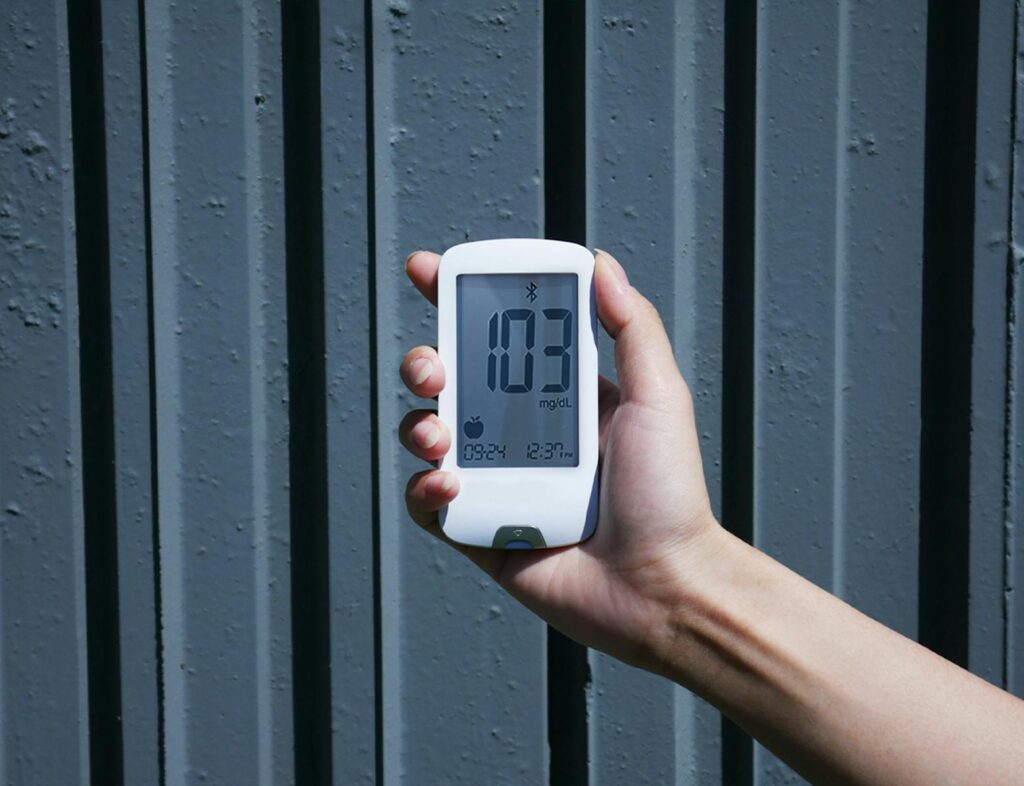 Close-up view of a hand holding a blood glucose meter displaying 103 mg/dL outdoors.