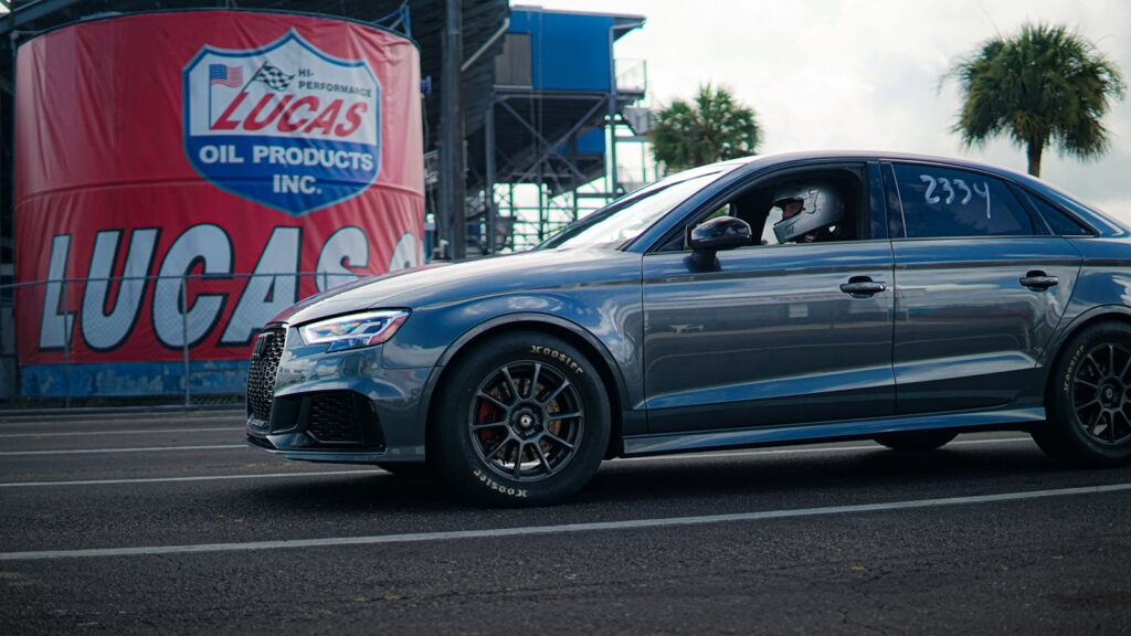 Gray performance car at Lucas Oil Raceway with driver wearing helmet.
