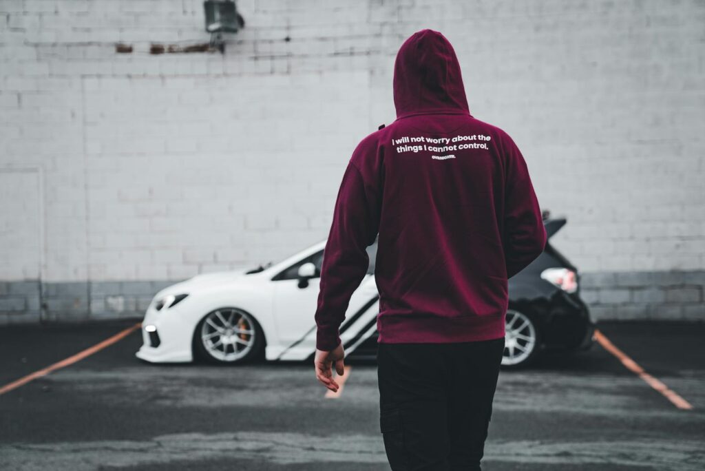 Back view of man in maroon hoodie walking towards two parked cars in a city parking lot.