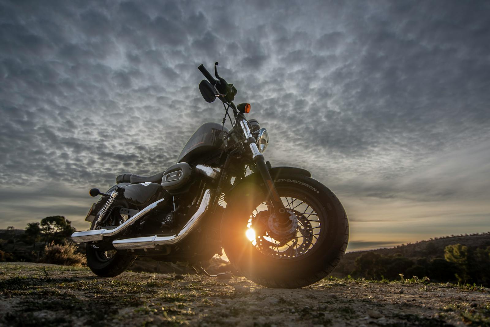 A striking image of a motorcycle parked on a dirt road with a dramatic sky at sunset, capturing the sun glare.