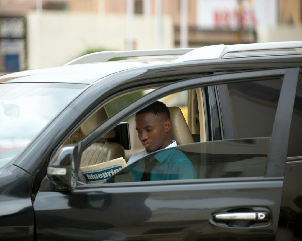 Man reading a newspaper inside his car, parked outdoors during the day.