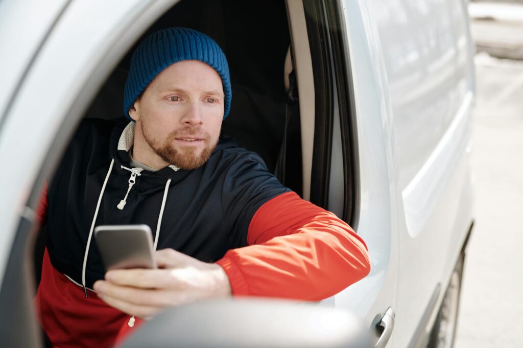 Stylish man in a car looking out the window holding a smartphone, wearing a beanie.