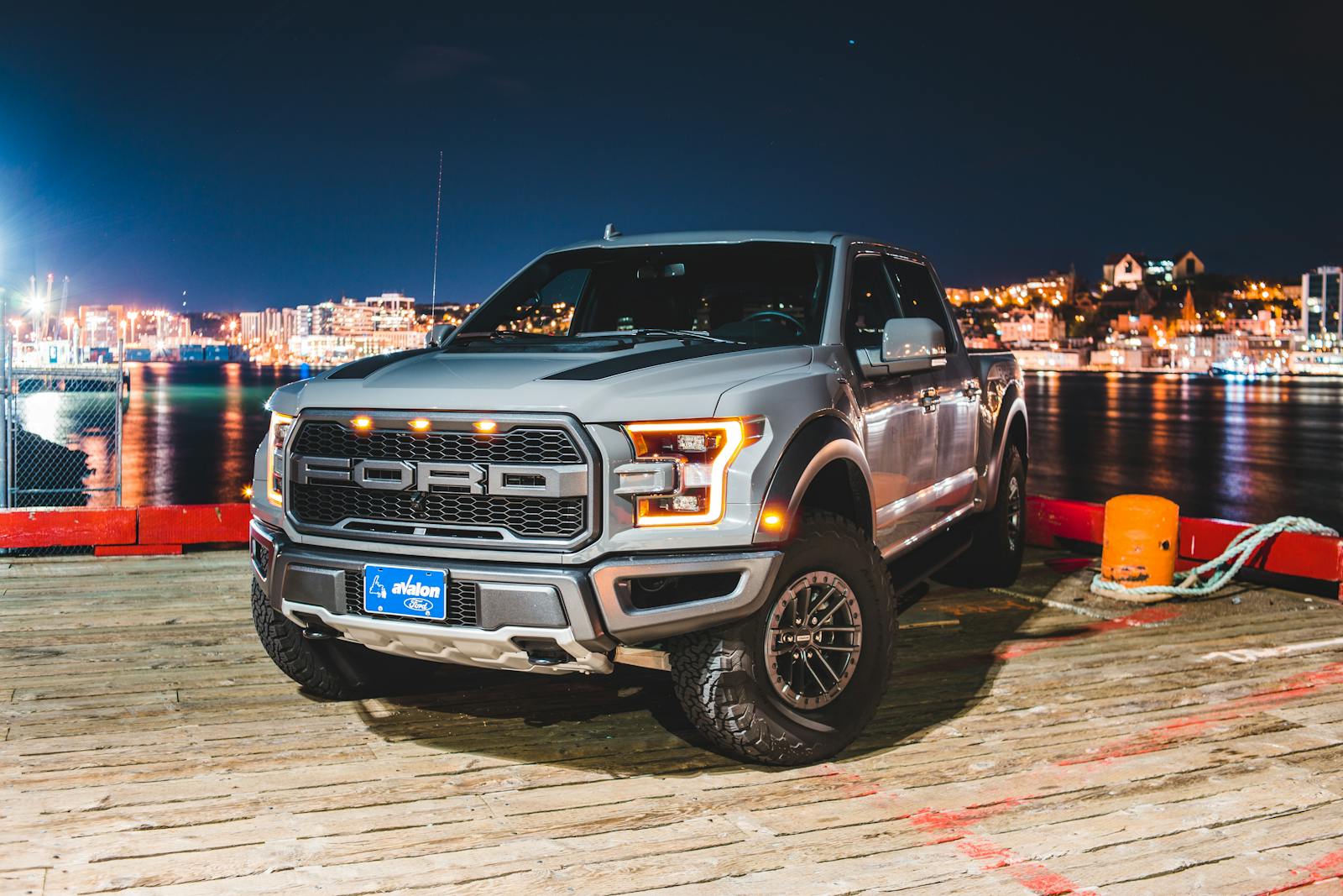 Silver pickup truck parked on a waterfront deck at night with city lights in the background.