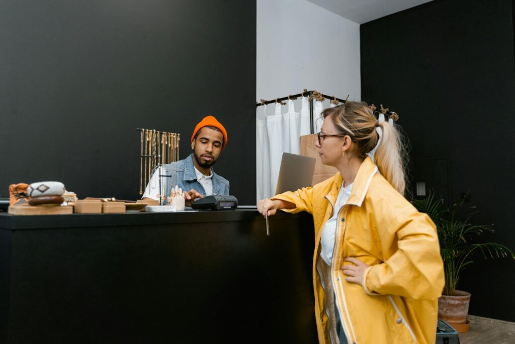 A woman pays with a card at a store counter to a bearded cashier in a denim vest.
