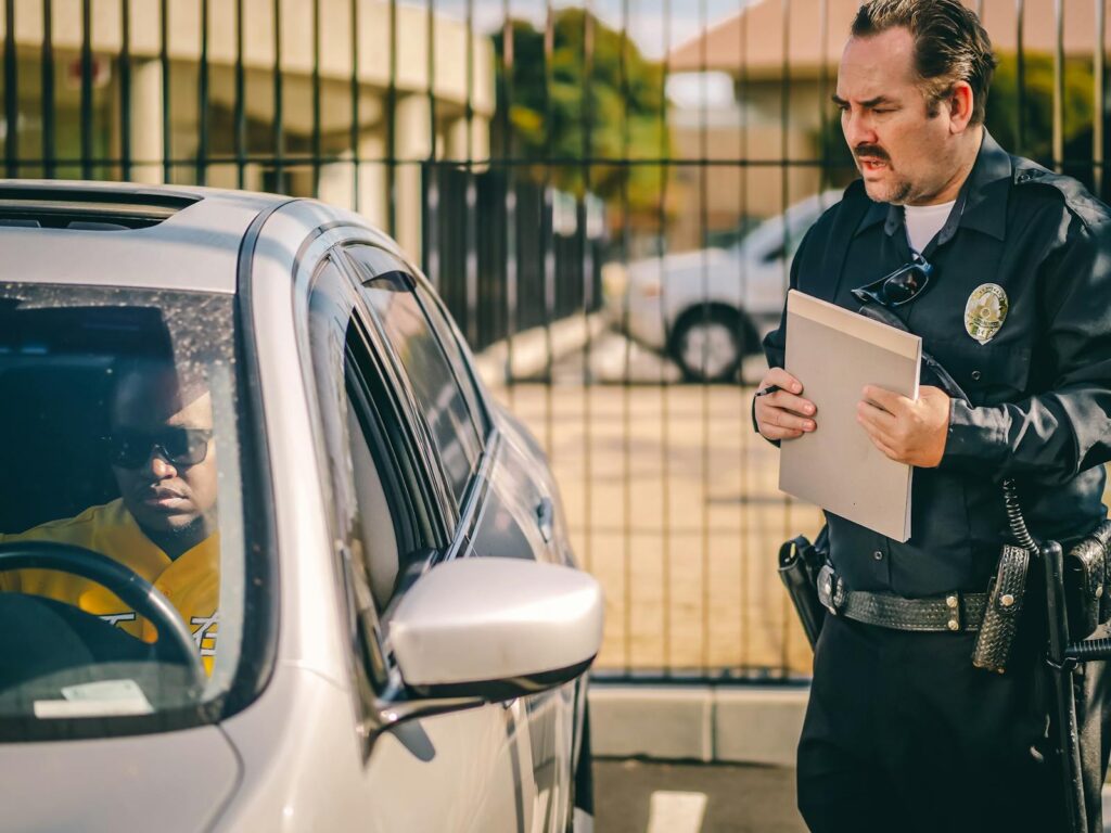 Police officer at car window issuing a ticket to a driver in a sunny outdoor setting.