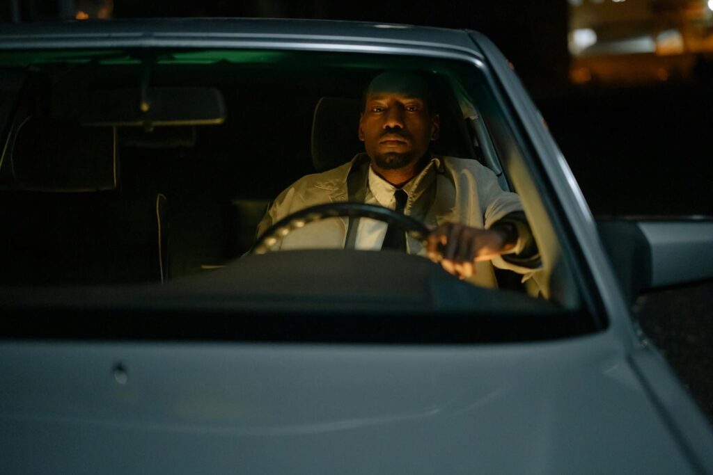 A man in a white jacket driving at night, focusing on the road ahead through the windshield.