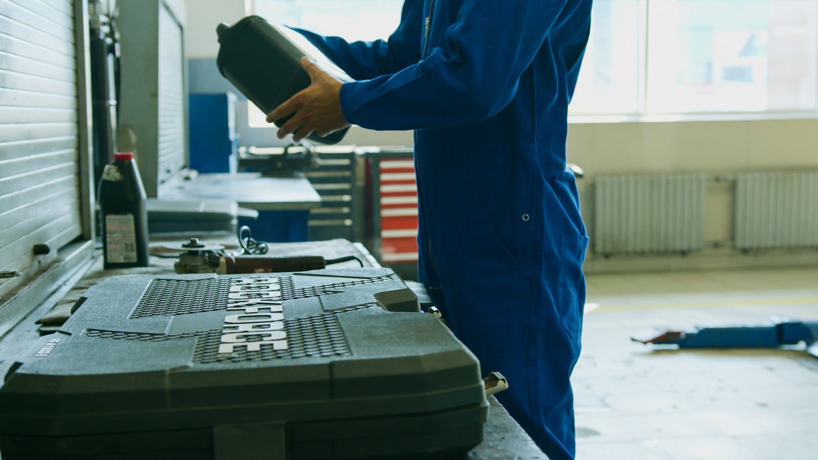 Mechanic in blue overalls holding engine oil container in a workshop environment, enhancing maintenance efficiency.
