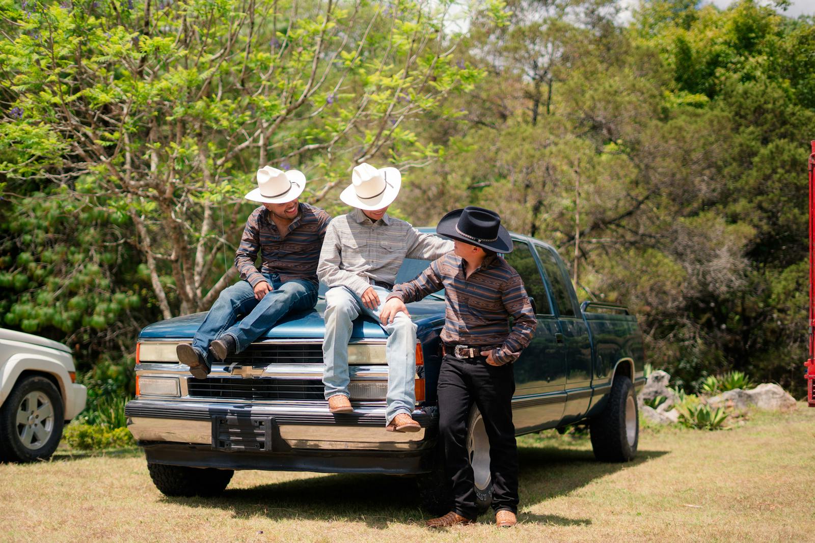 Three cowboys in hats relaxing on a truck in a sunny countryside setting.