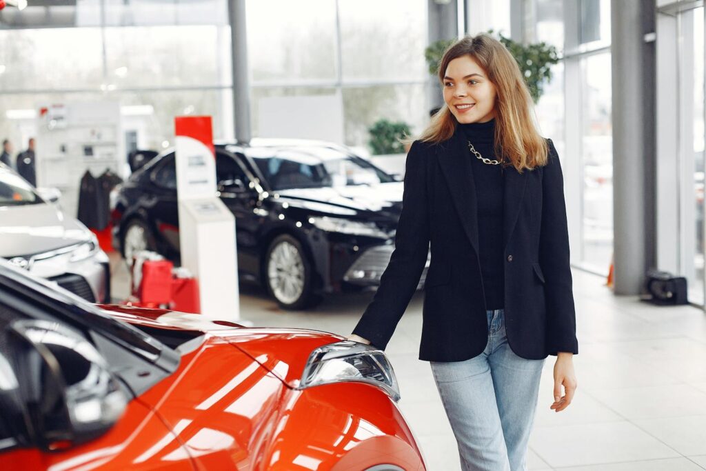 Smiling woman exploring cars in a bright showroom, expressing enthusiasm.