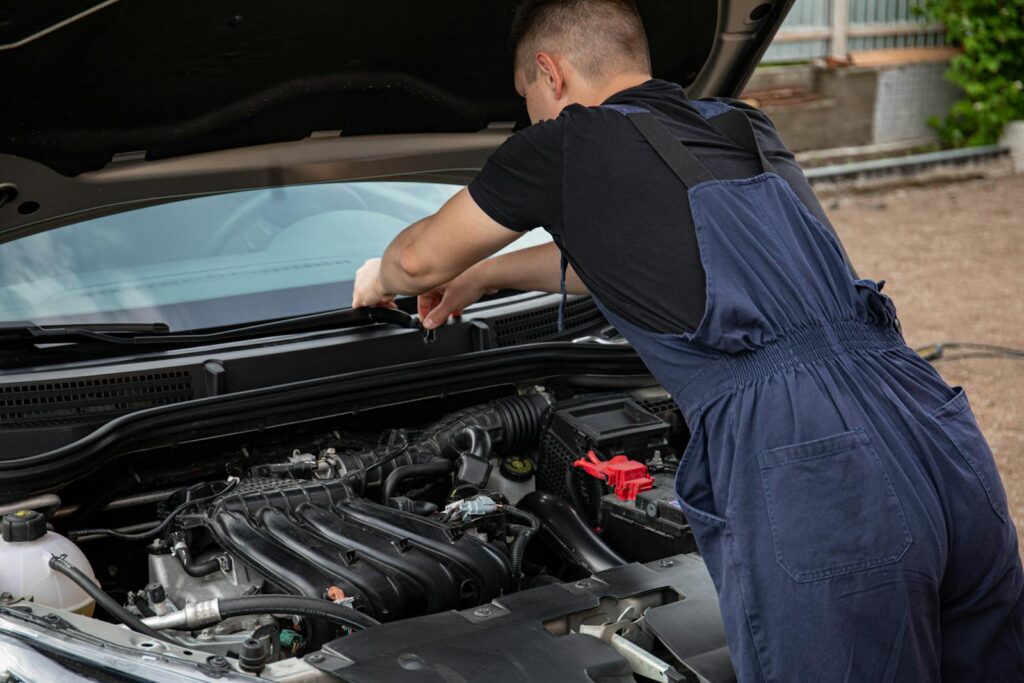 A mechanic in blue overalls works on a car engine outdoors, showcasing vehicle maintenance skills.
