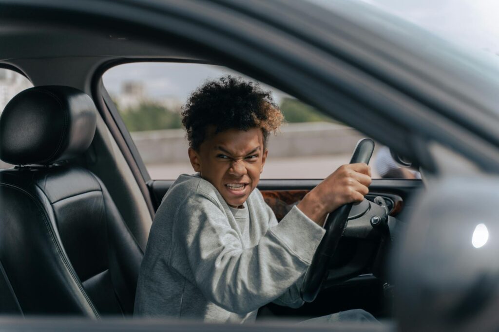 A teenager in a car momentarily pretending to drive with a determined expression while sitting indoors.