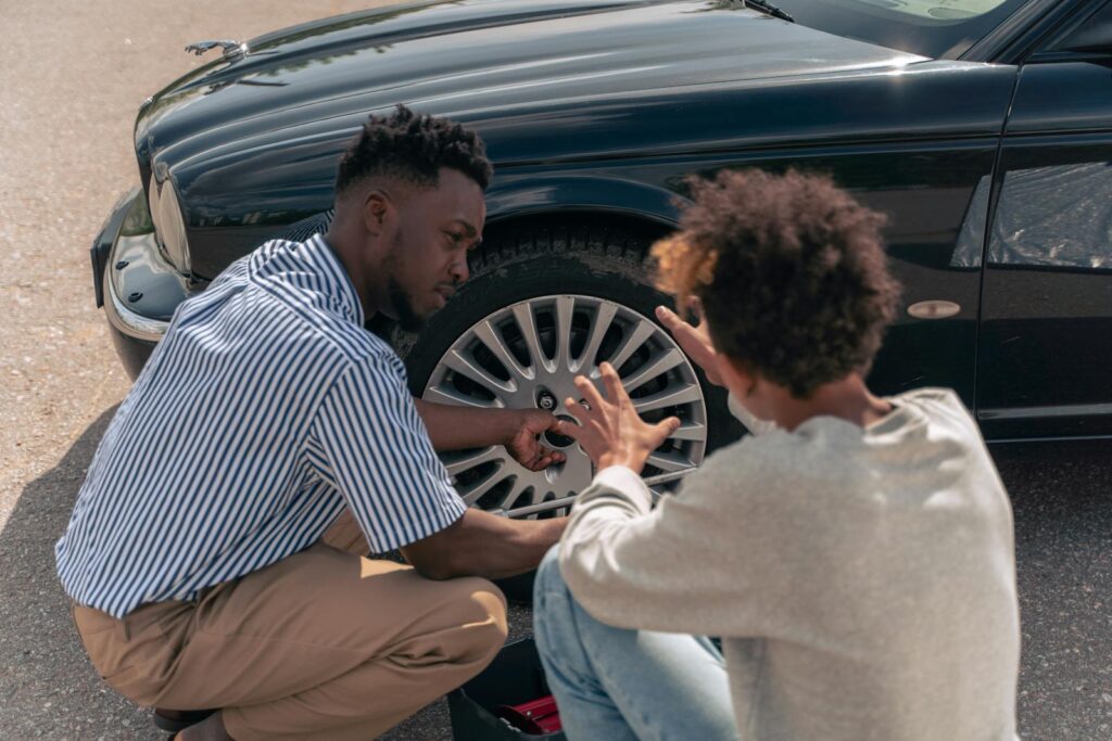 A father instructs his teenage son on how to change a car tire outdoors.