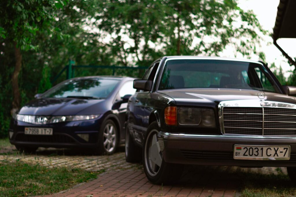 A classic car and a modern car parked in a shaded driveway, surrounded by greenery.