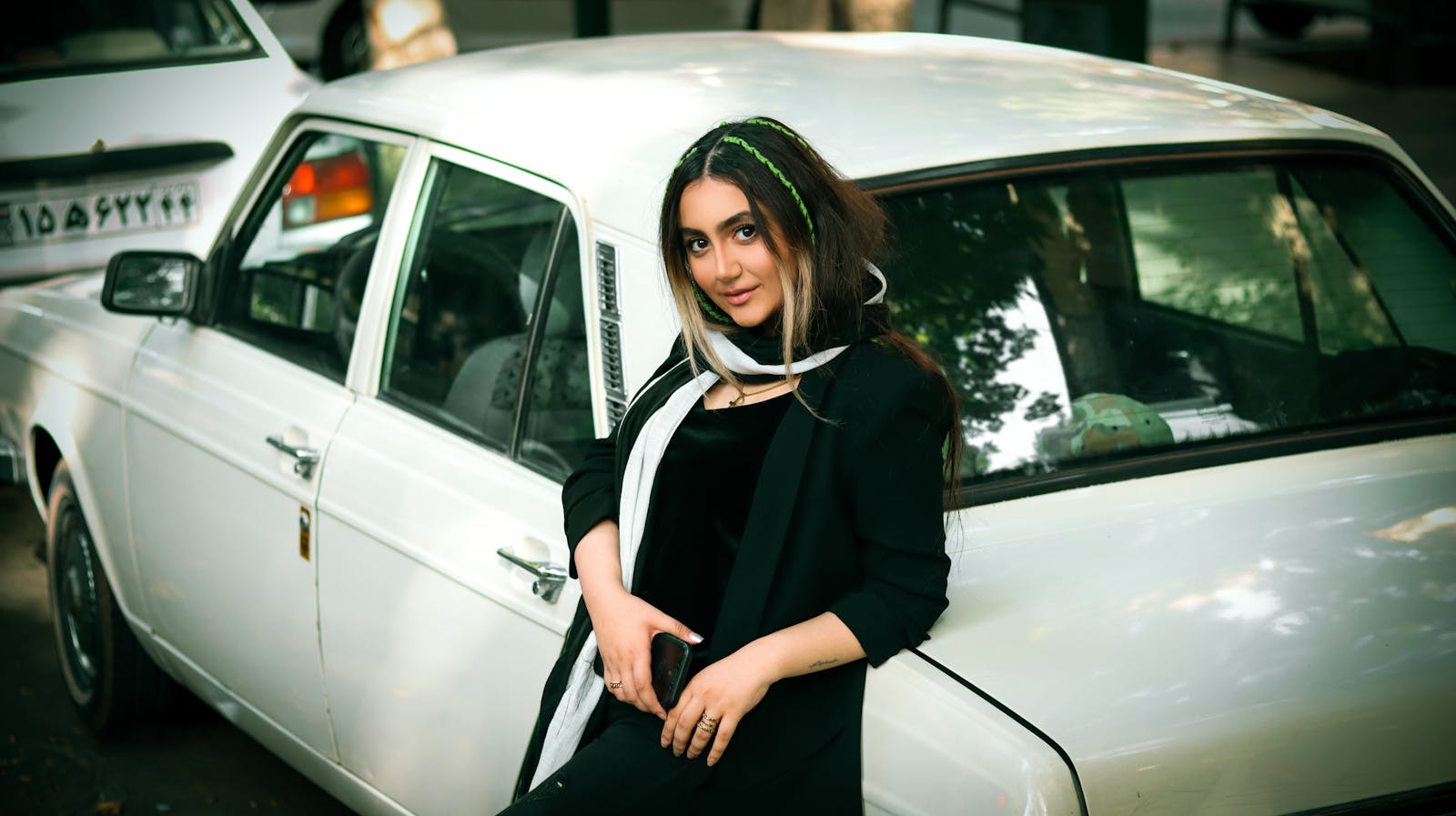 Fashionable woman leaning by a vintage car on a Tehran street, exuding urban elegance.