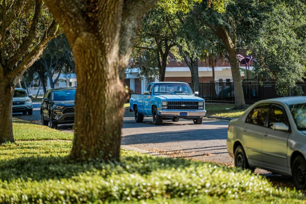 A classic blue pickup truck drives down a sunny, tree-lined suburban street.