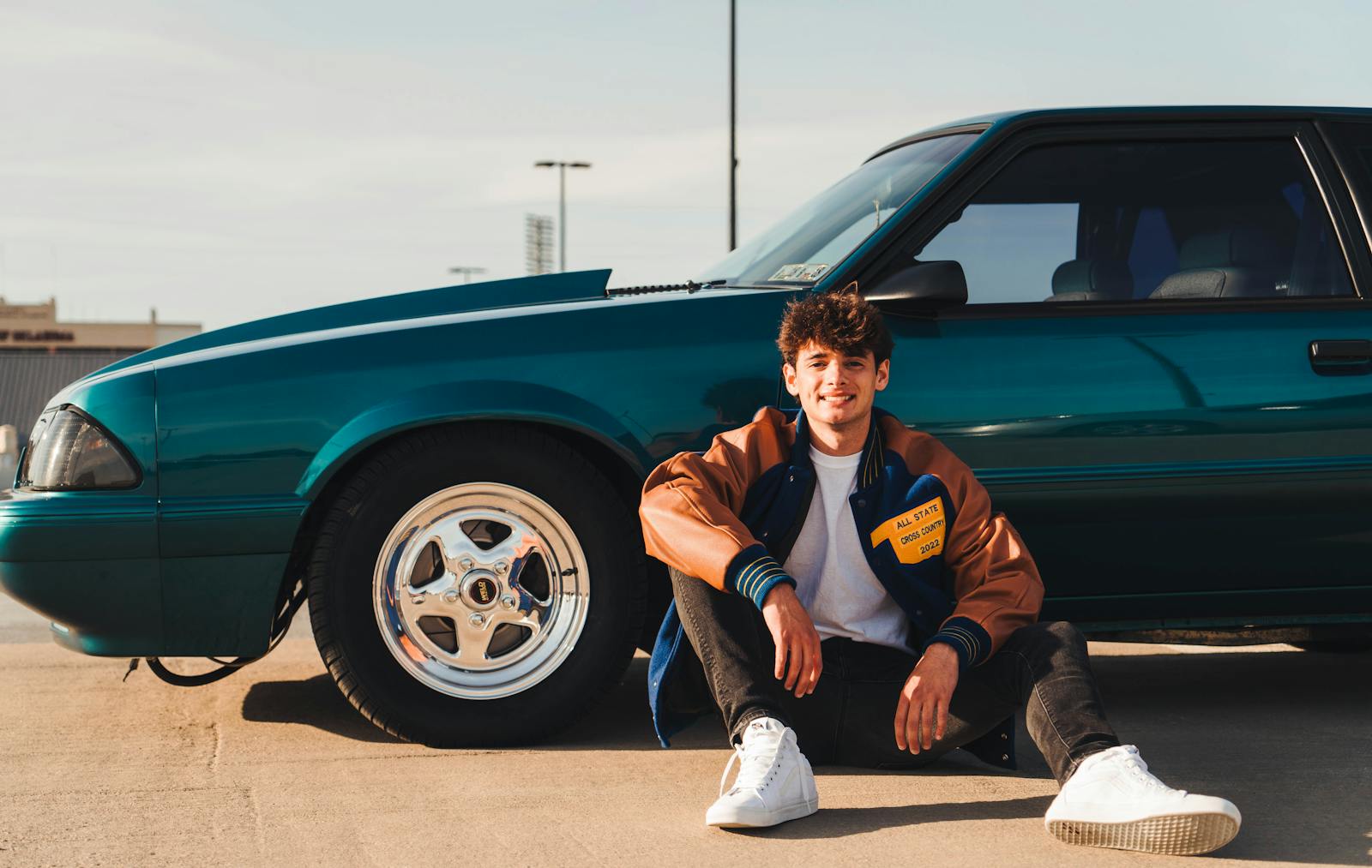 Man sitting by a teal car, wearing a varsity jacket, in Norman, Oklahoma.