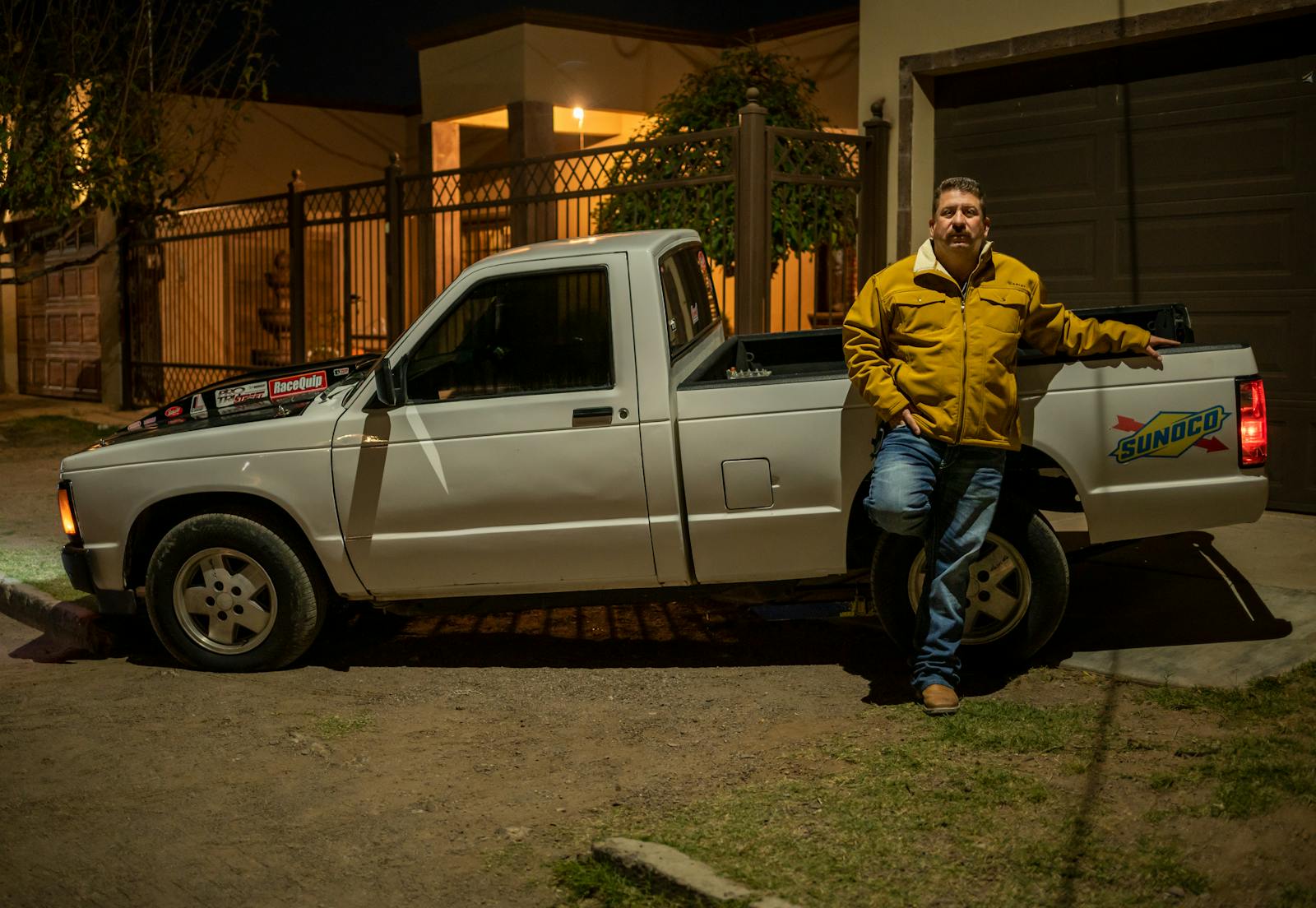 A man in a yellow jacket leaning on a white pickup truck at night in an urban area.