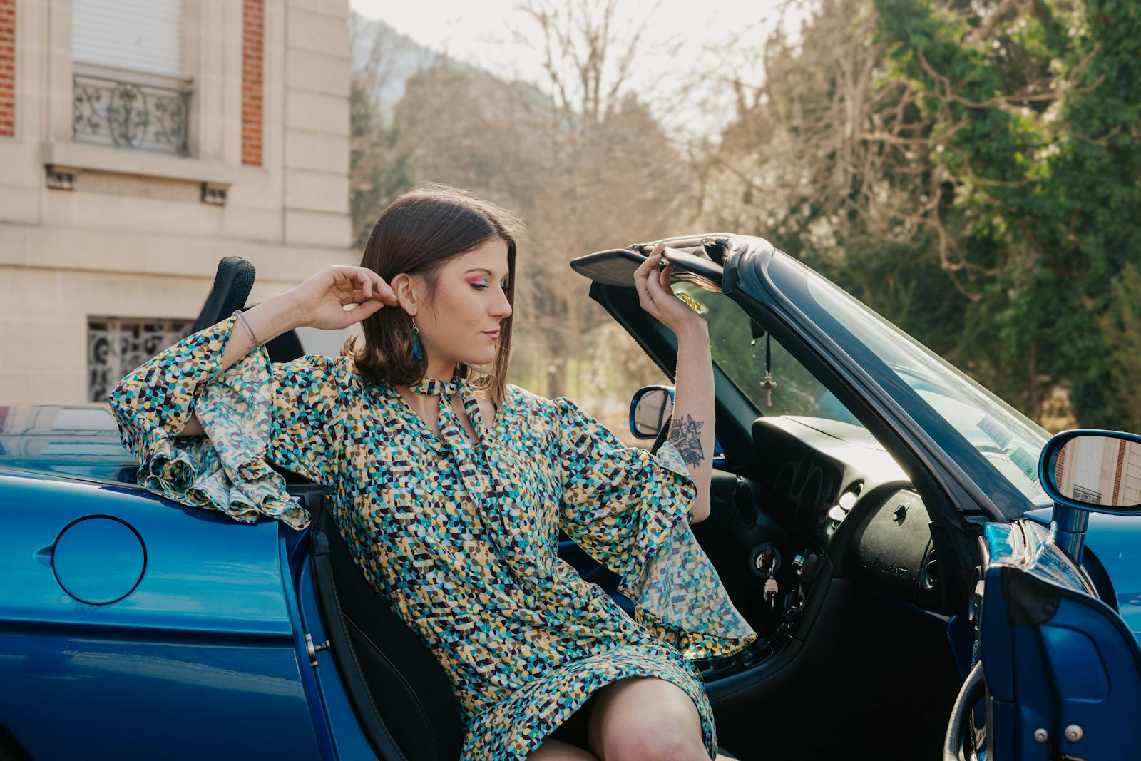 Stylish woman in a floral dress sits in a blue convertible car on a sunny day.