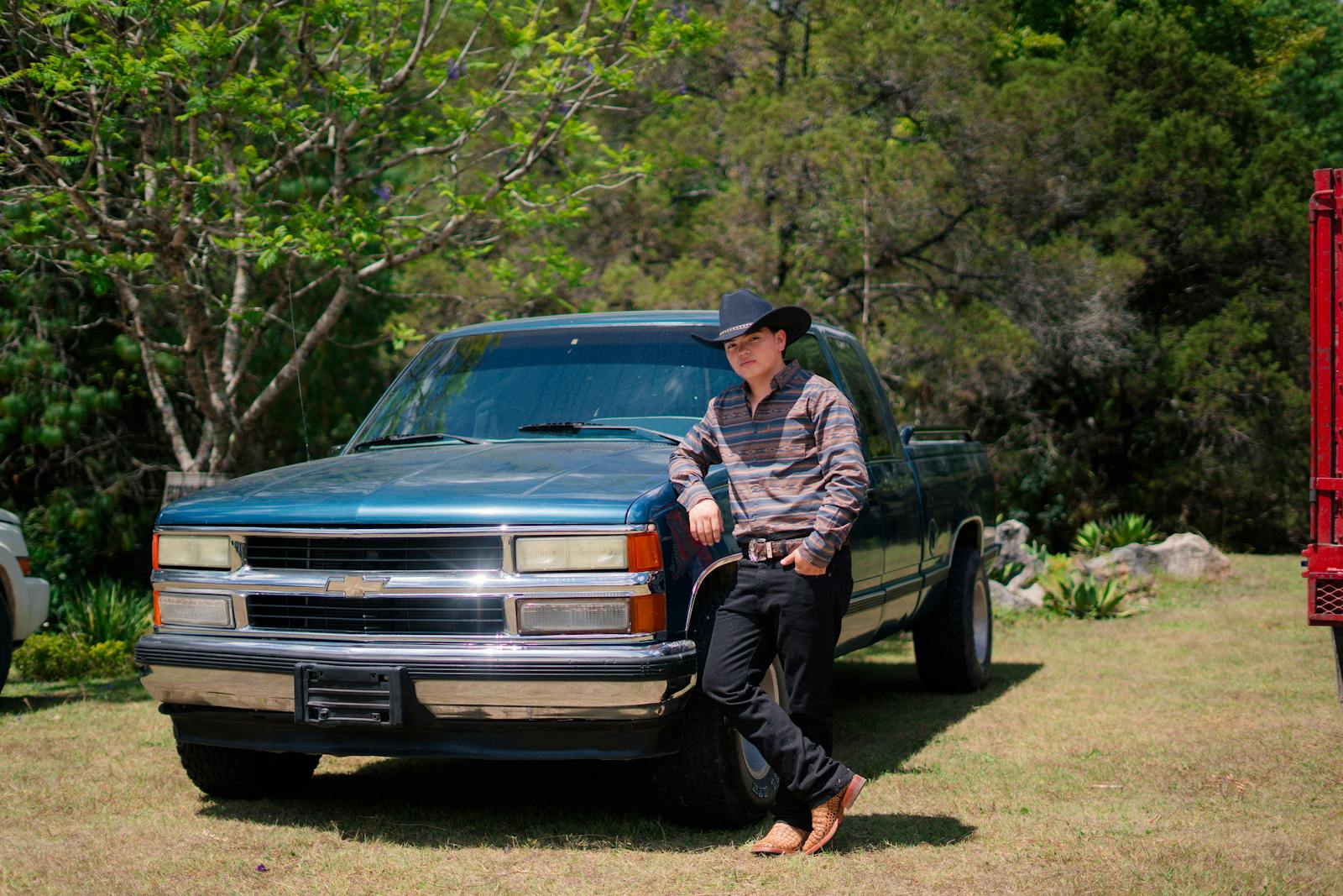 A cowboy in a hat relaxes against a classic blue truck outdoors in a sunny field.