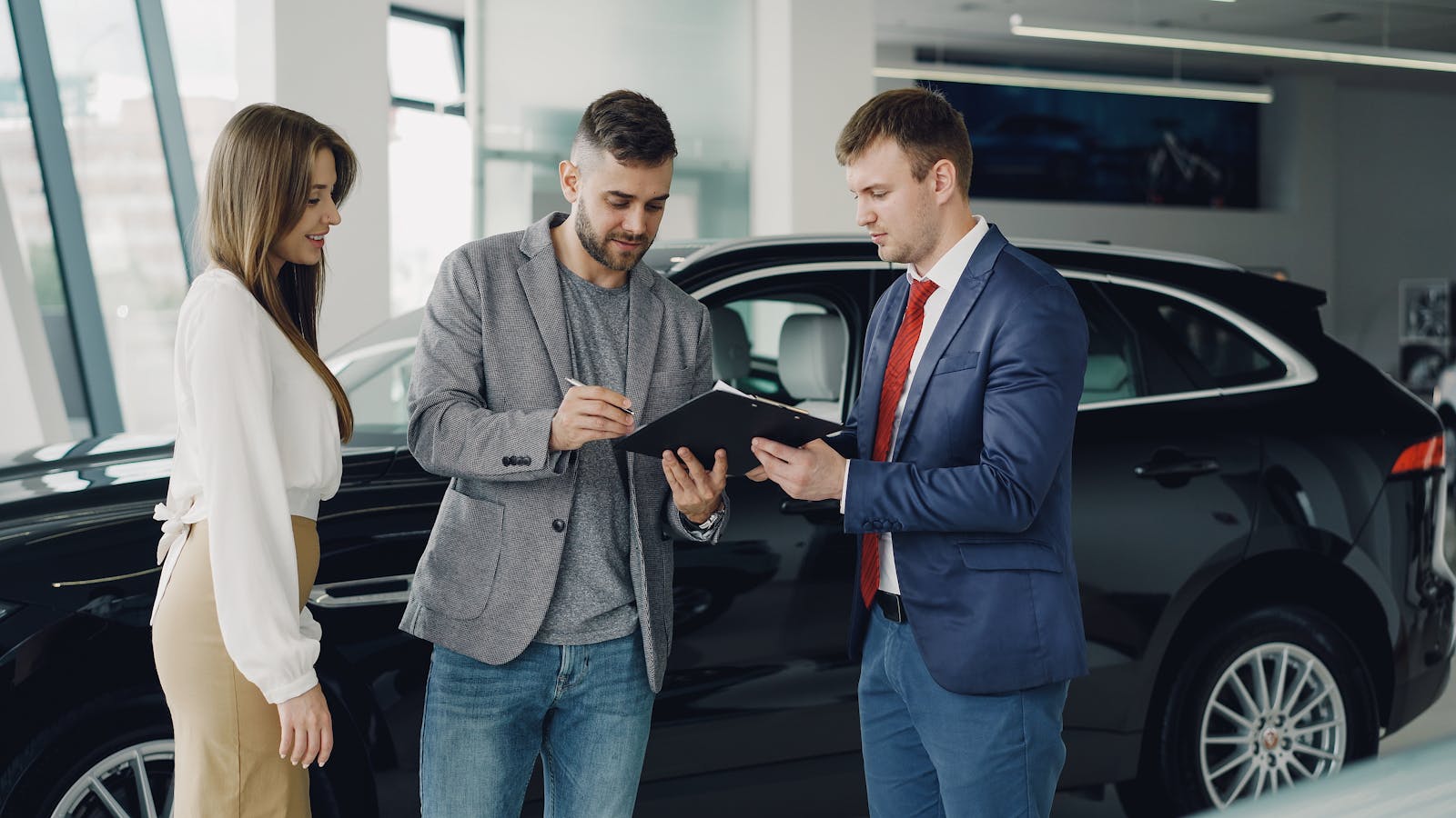 Three adults discussing documents at a car dealership beside a black car.