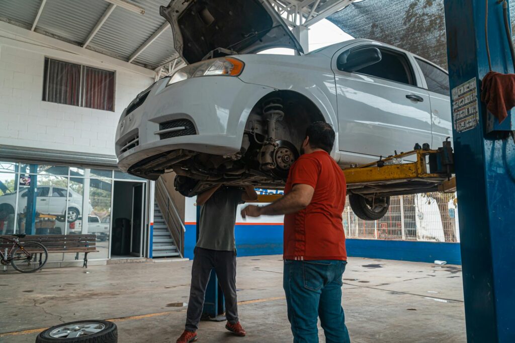Two mechanics inspecting a white car on a lift inside a garage workshop.