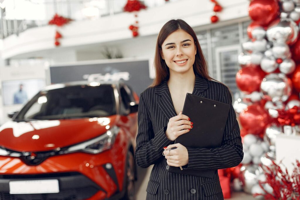 Cheerful friendly stylish female consultant in trendy black formal dress standing with clipboard standing in car showroom against new red shiny car and looking at camera