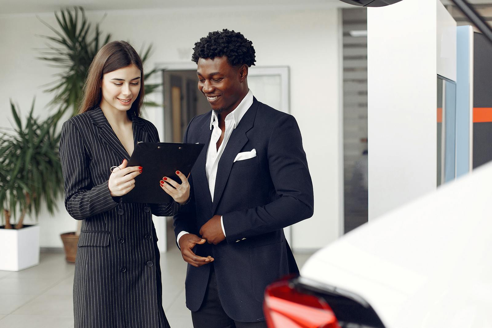 Cheerful multiethnic stylish female agent and smiling customer in formal suit discussing contract details while standing in modern car showroom in daylight
